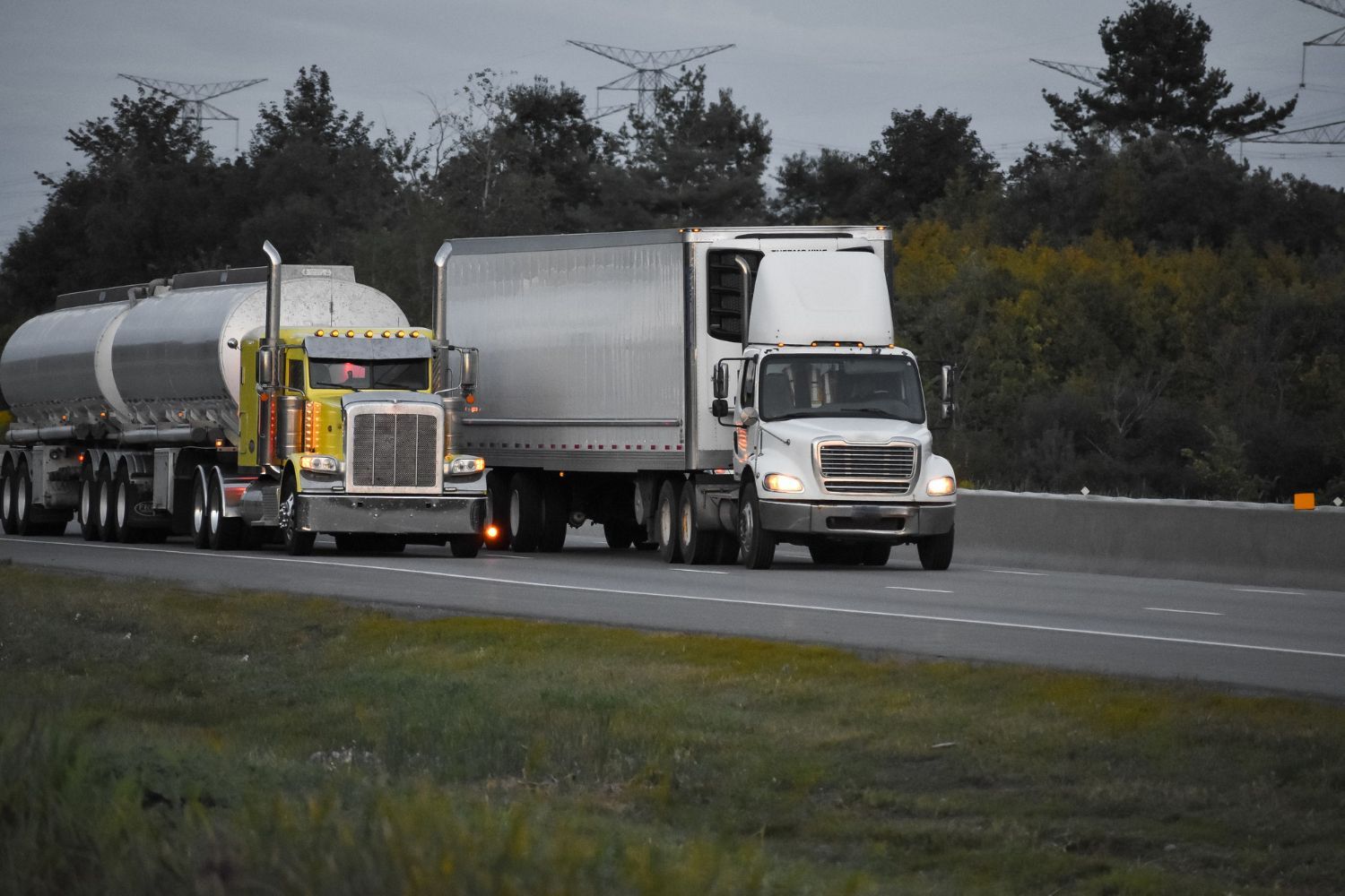 Two semi trucks driving side by side on a highway, one hauling a silver tanker trailer.