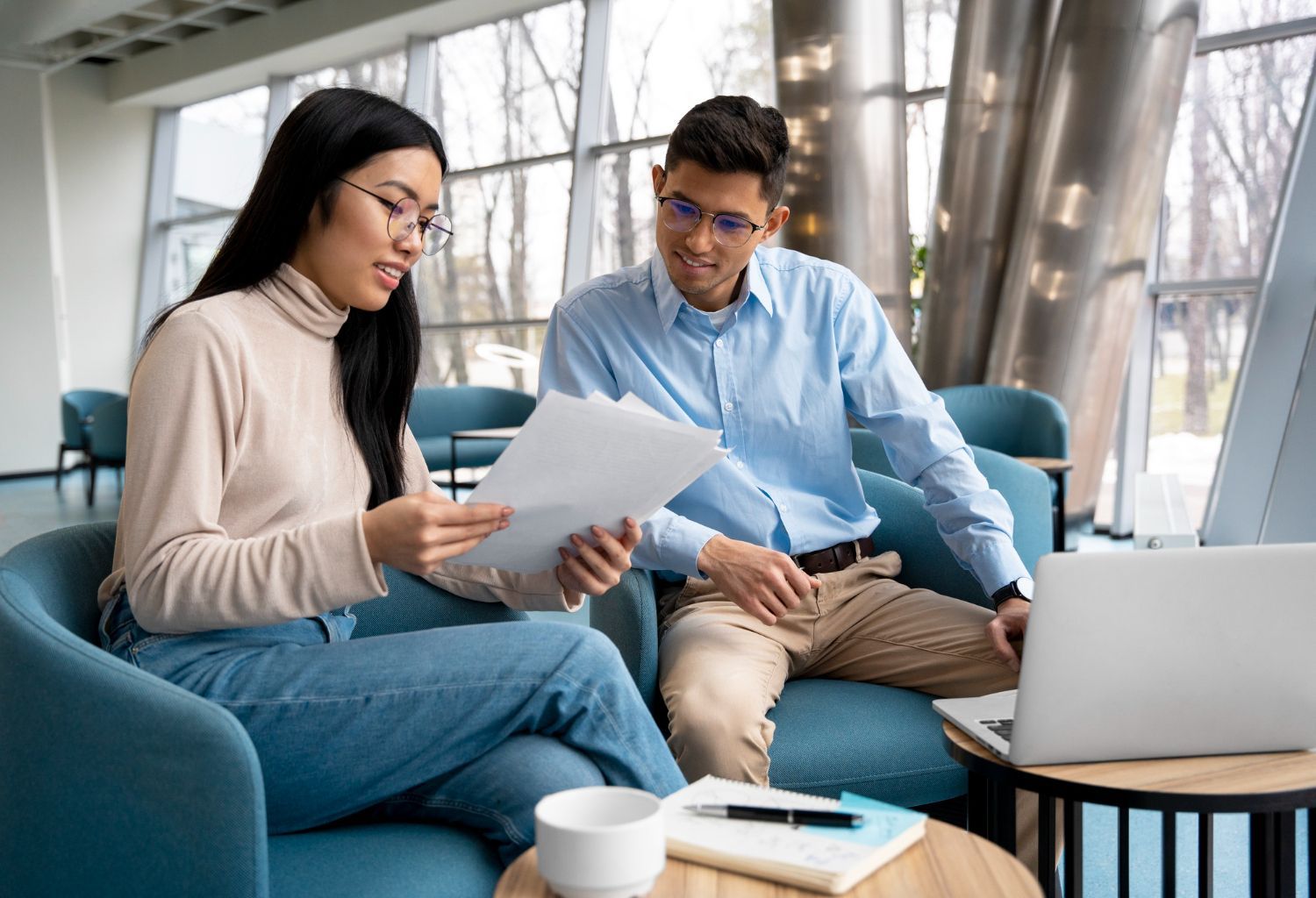 Two coworkers reviewing papers on a sofa beside a laptop in a bright office lounge