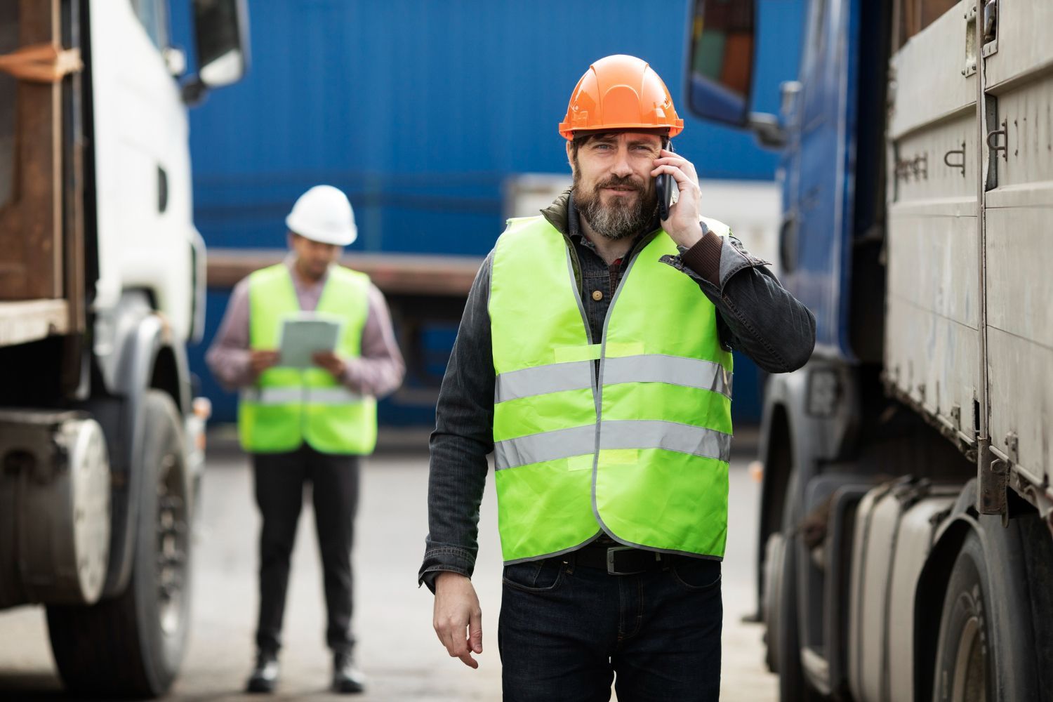 Construction workers in hard hats and safety vests between trucks, one speaking on a phone