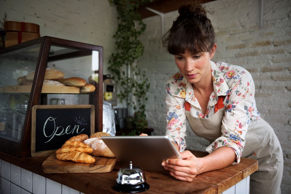 Bakery counter with an apron-clad worker using a tablet beside a chalkboard “Open” sign and croissants
