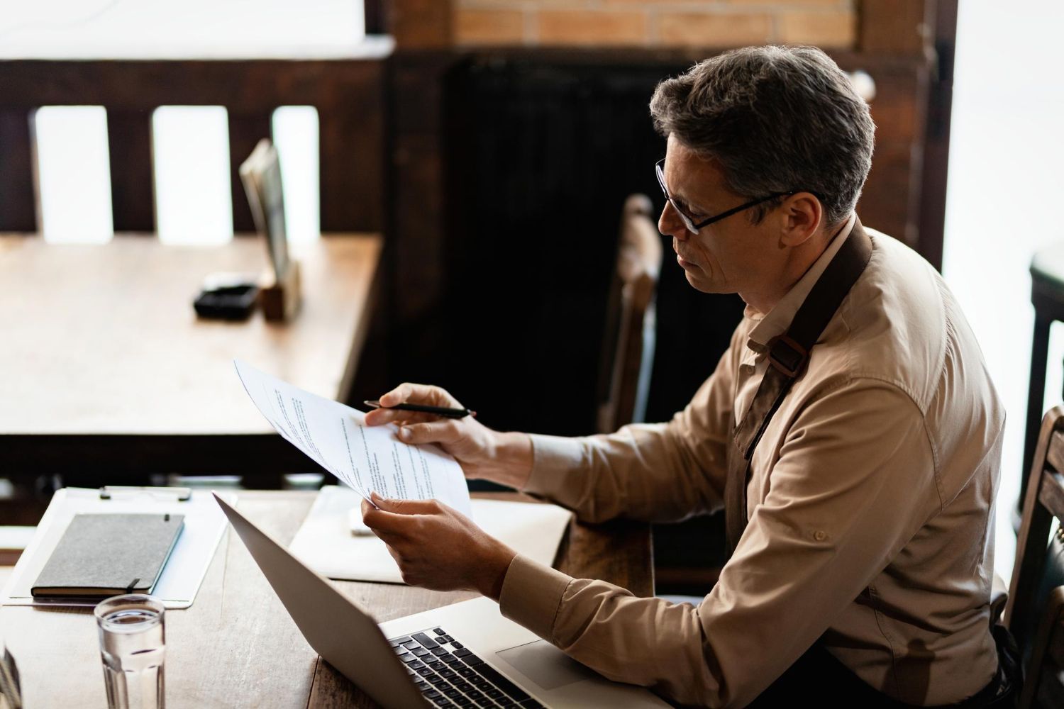 Man seated at a table using a laptop and holding a document in a bright office setting