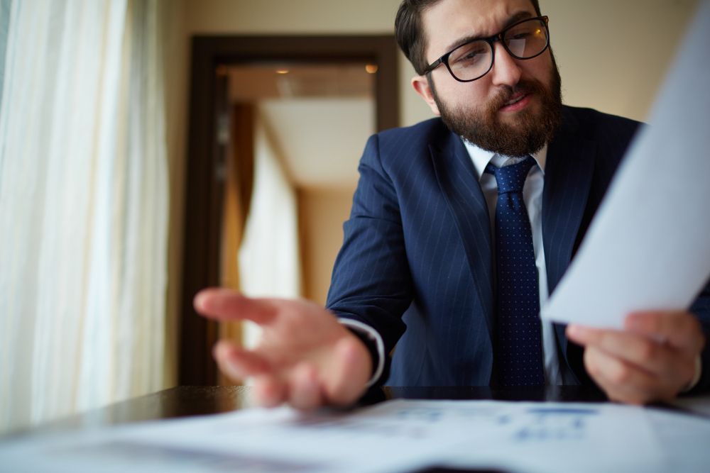 Man in a blue suit and glasses reviewing papers at a desk in a hallway