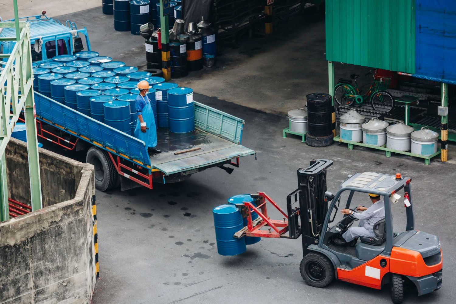 Orange forklift moving a blue barrel beside a flatbed truck loaded with blue drums in an industrial yard