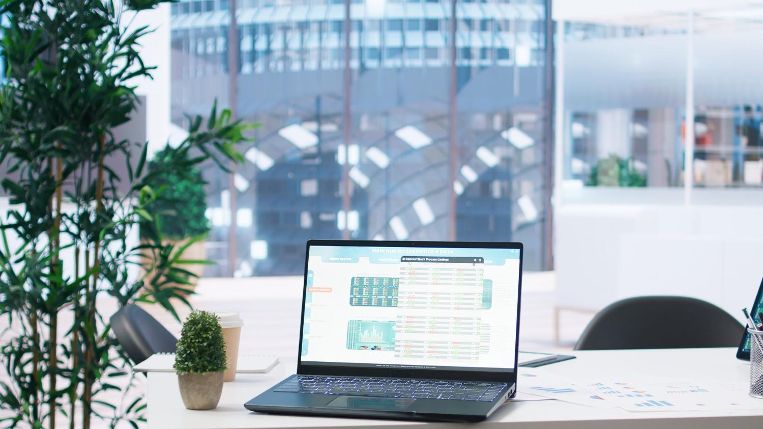 Laptop on a bright office desk beside a potted plant, with a blurred city view in the background.