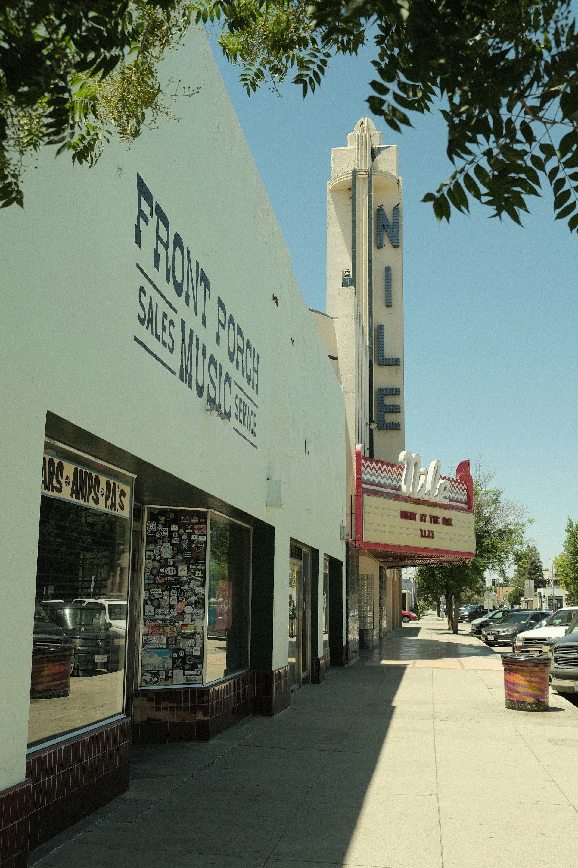 Sunny sidewalk beside a theater building with a marquee and storefronts under trees