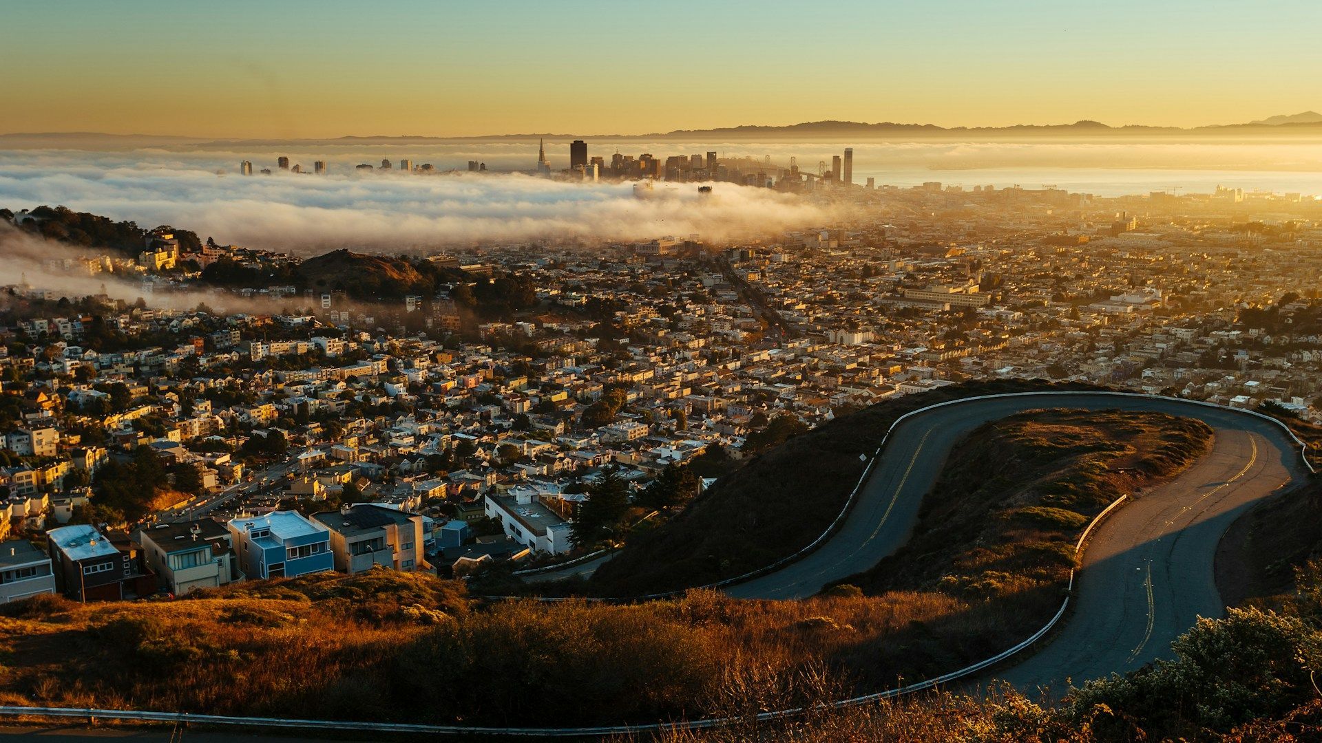 Golden sunrise over a sprawling city and winding hillside road, with fog blanketing the distant skyline