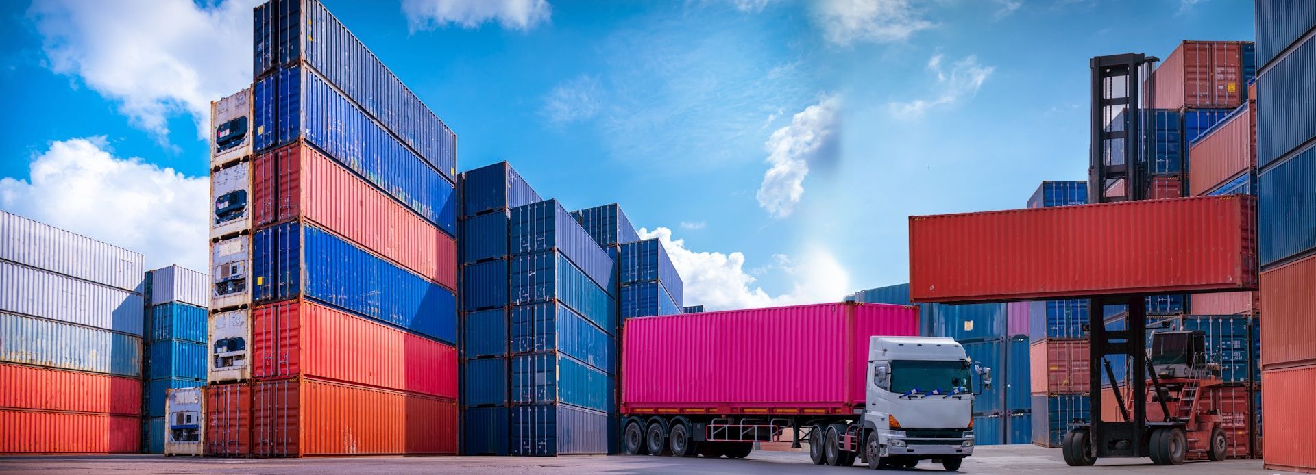 Colorful shipping containers stacked in a port yard with a truck under a blue sky.