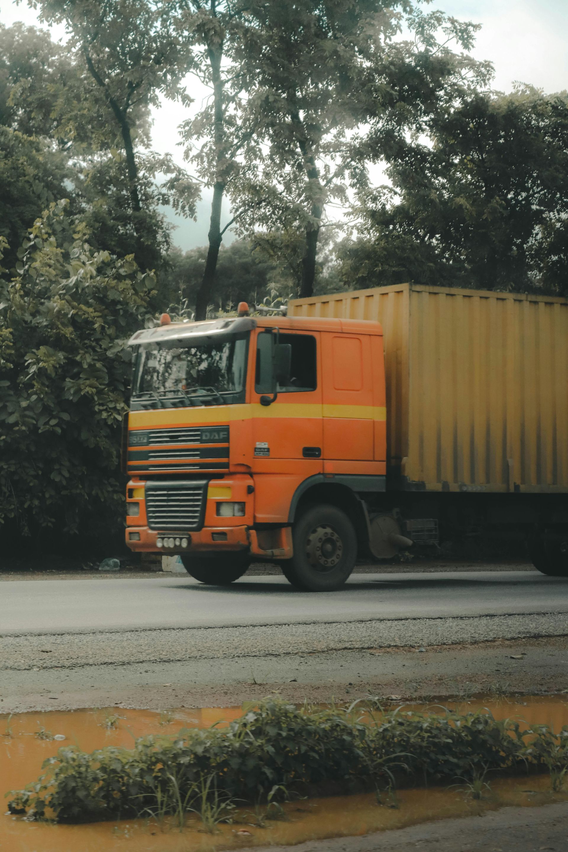 Orange cargo truck driving on a road beside trees and a median strip.