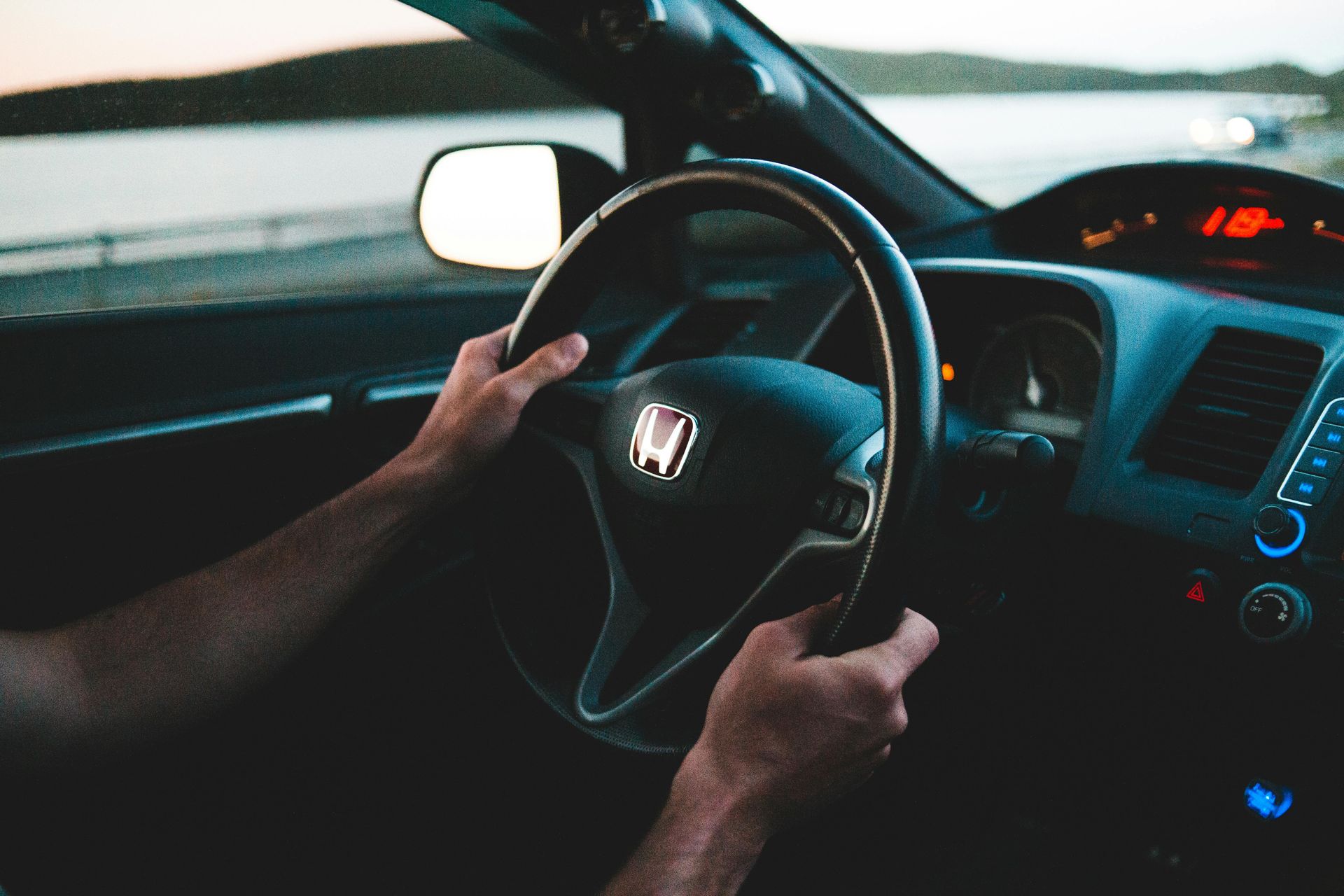 Driver’s hands on a Honda steering wheel inside a car, dashboard lights glowing at dusk.