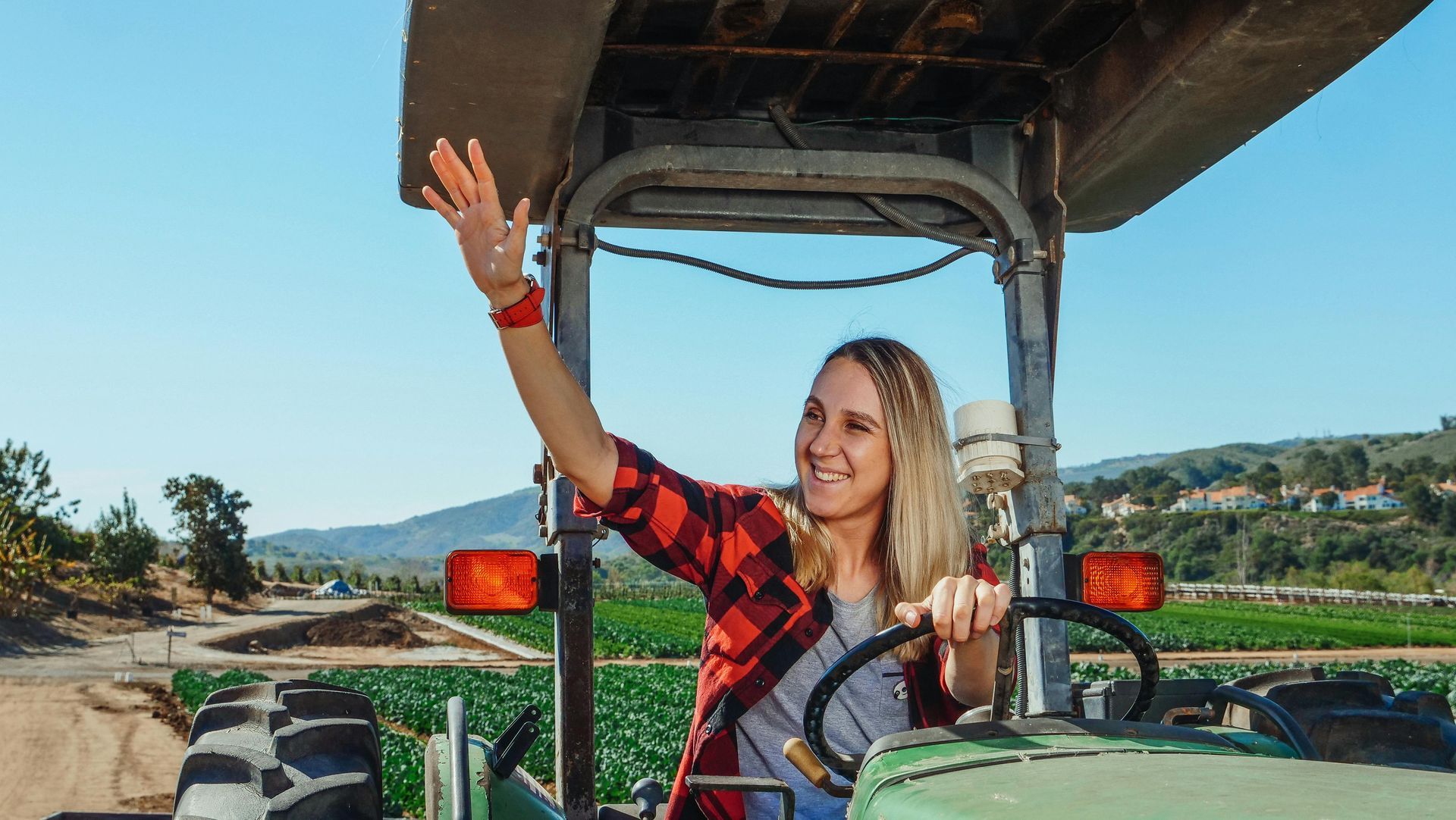 Person in plaid shirt waves from an open safari vehicle on a sunny road.