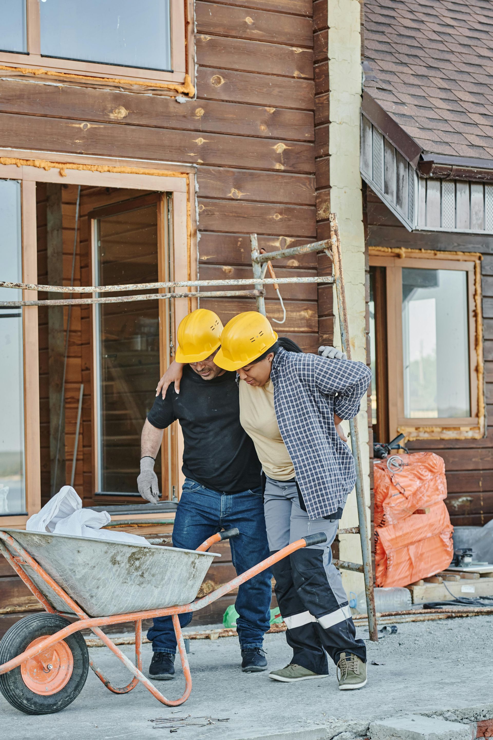 Two workers in yellow hard hats lean over a wheelbarrow outside a wooden building.