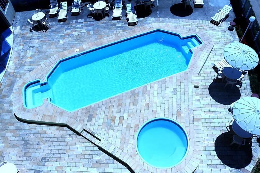 Vista aérea de uma piscina azul-clara e uma área de spa em um pátio de pedra bege, com espreguiçadeiras e guarda-sóis