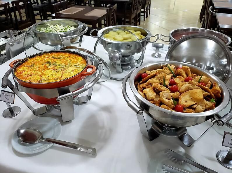 Mesa de bufê com várias réchauds com pratos quentes variados e uma colher de servir