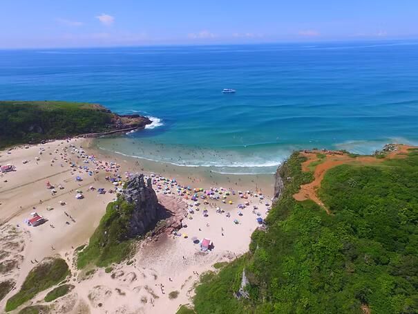 Vista aérea de uma praia arenosa lotada, com águas azul-turquesa, falésias verdes e uma pequena enseada