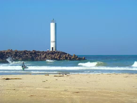 Praia com um farol branco sobre um molhe rochoso sob um céu azul e limpo