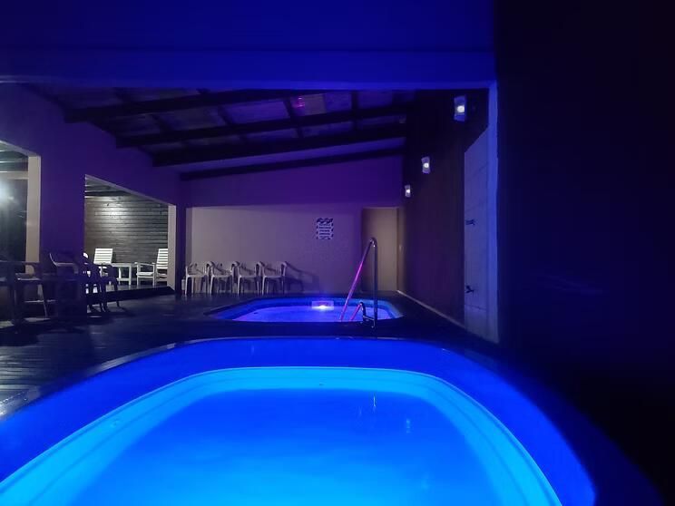 Piscina coberta iluminada em azul à noite, com espreguiçadeiras e uma área de pátio coberta