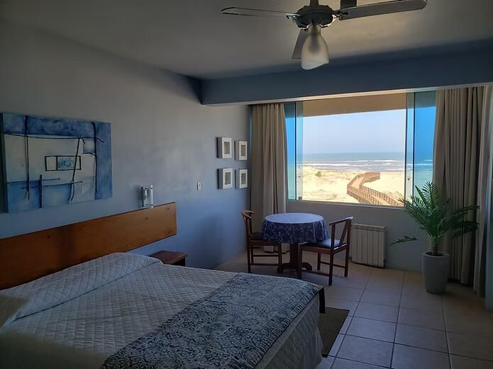 Quarto com vista para o mar, cama, mesinha e portas da varanda abertas com vista para a praia