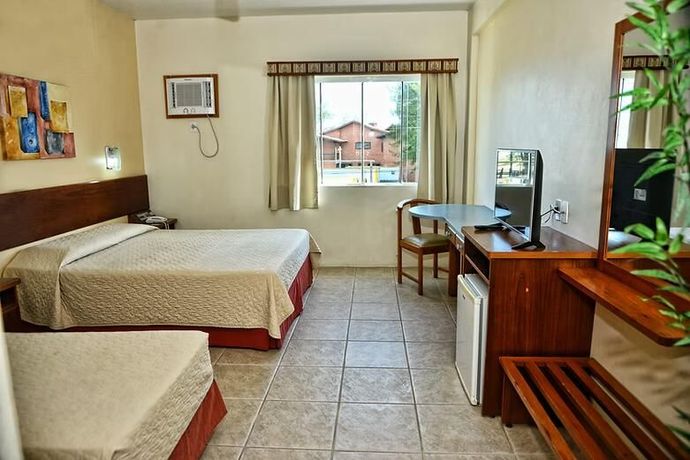 Quarto de hotel com duas camas, piso de azulejos, escrivaninha, espelho e uma janela com cortinas