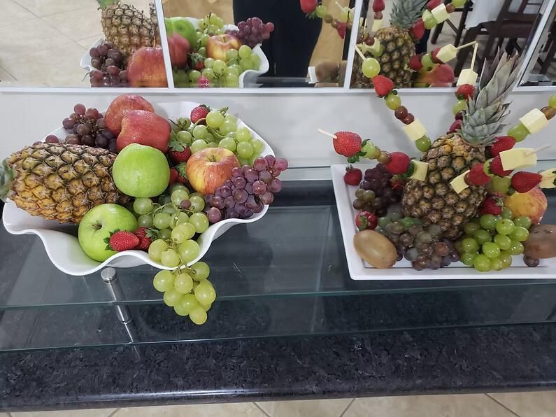 Travessas decorativas de frutas com uvas, maçãs, abacaxis e morangos sobre um balcão de vidro.