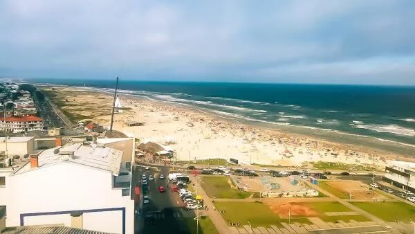 Vista de uma praia e do oceano numa cidade costeira, sob um céu nublado, vista de cima, com uma estrada e edifícios em primeiro plano