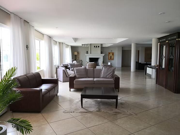 Sala de estar moderna com sofá de couro marrom, assentos modulares, mesa de centro e grandes janelas com cortinas translúcidas