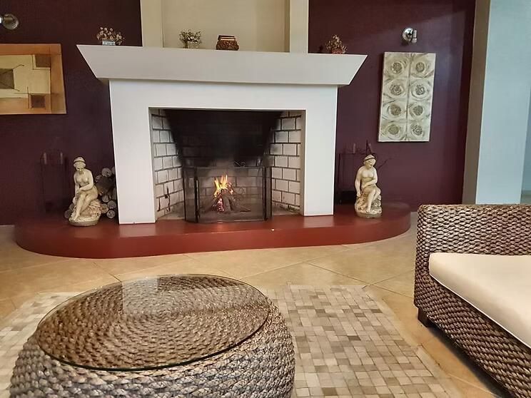 Sala de estar com lareira branca, assentos de vime e piso de mosaico, iluminada por uma pequena fogueira
