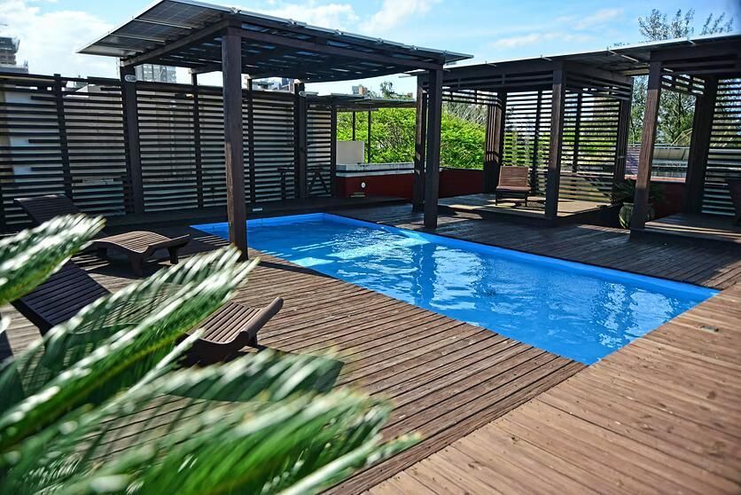 Deck de madeira coberto, com uma piscina azul e uma pérgola com ripas sob a luz intensa do sol
