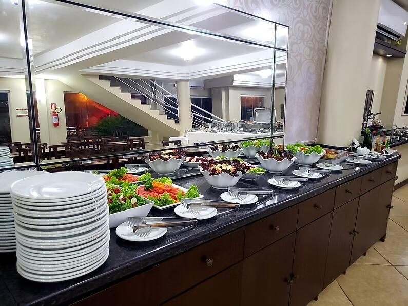 Mesa de buffet com pratos brancos empilhados, saladas e travessas numa sala de refeições bem iluminada