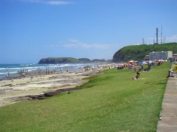 Praia ensolarada com passeio relvado, ondas e pessoas a caminhar à beira-mar