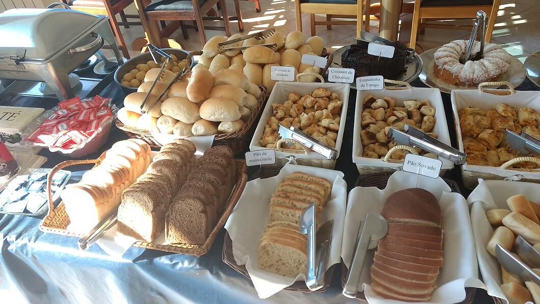 Pães e doces variados dispostos em bandejas sobre uma mesa de buffet com toalha branca