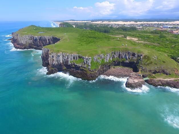 Falésias costeiras verdes sobre o oceano turquesa, com ondas brancas e uma costa distante sob o céu azul