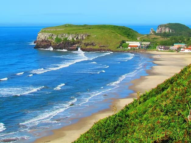 Praia de areia com ondas azuis, falésias verdes e um promontório relvado sob um céu limpo.