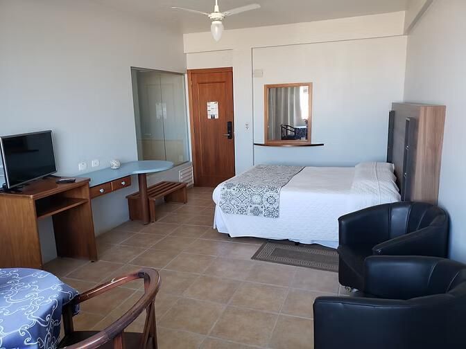 Quarto de hotel simples com duas camas de solteiro, TV, escrivaninha, cadeira, piso de cerâmica e espelho na parede