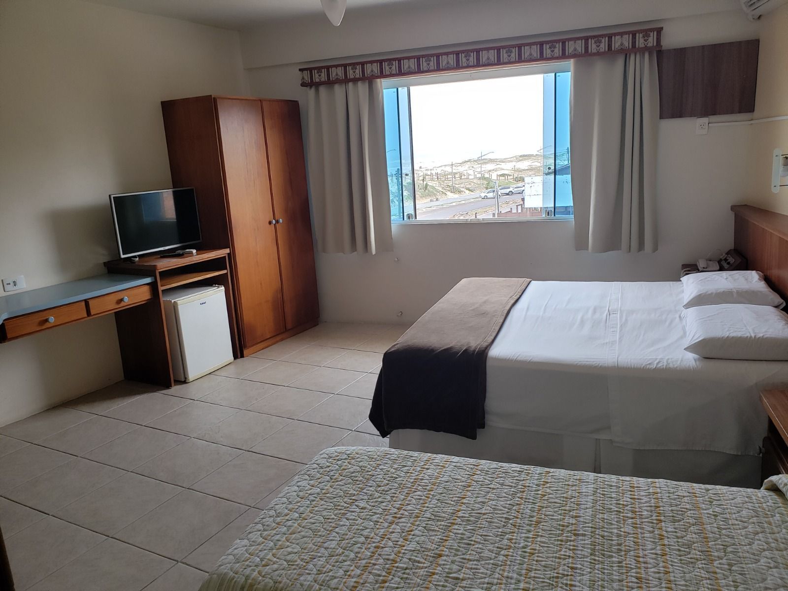Quarto de hotel com duas camas, escrivaninha, guarda-roupa e uma janela com vista para a cidade.