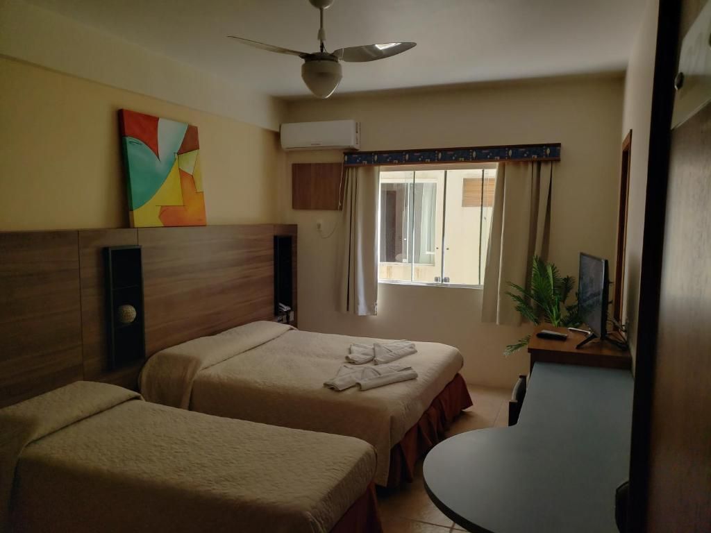 Quarto pequeno com duas camas, parede revestida de madeira, ventilador de teto, janela e uma planta ao lado da cômoda