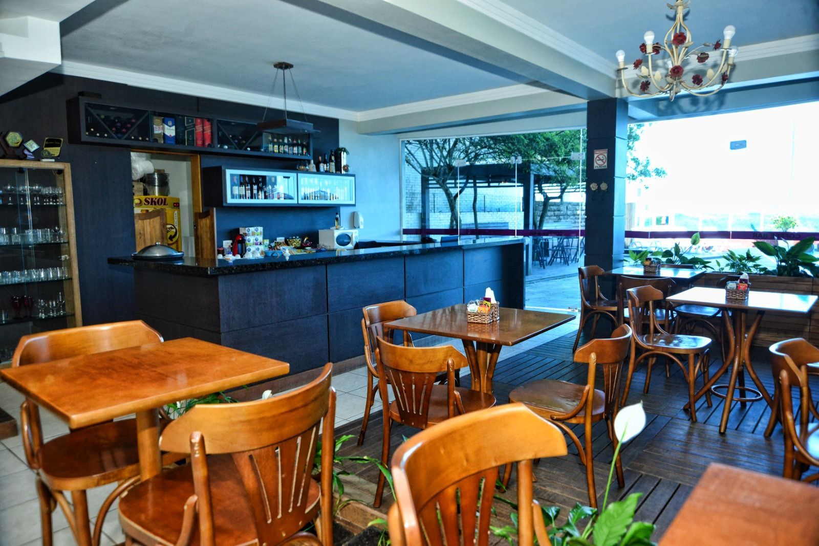 Interior aconchegante de café, com mesas e cadeiras de madeira, balcão escuro e grandes janelas luminosas