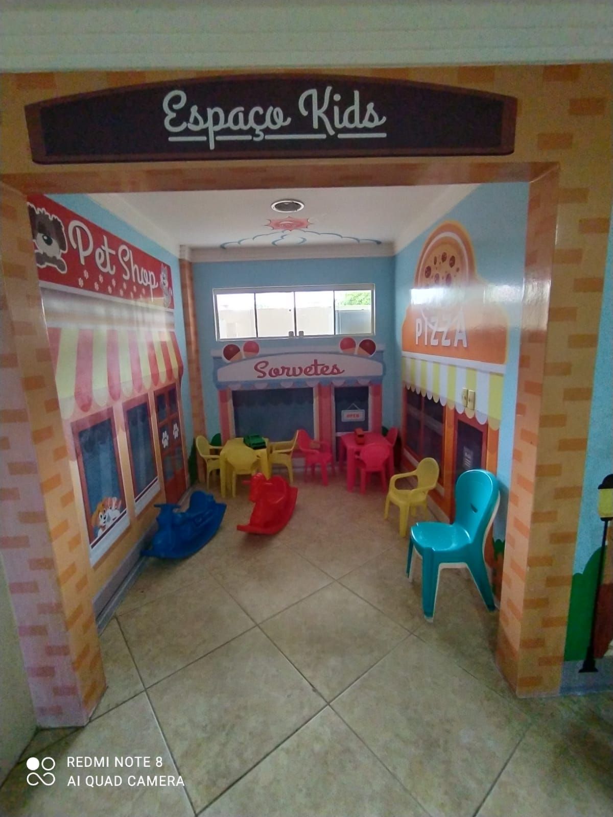 Área infantil com cadeiras de plástico coloridas e uma pequena barraca dentro de uma sala revestida de azulejos