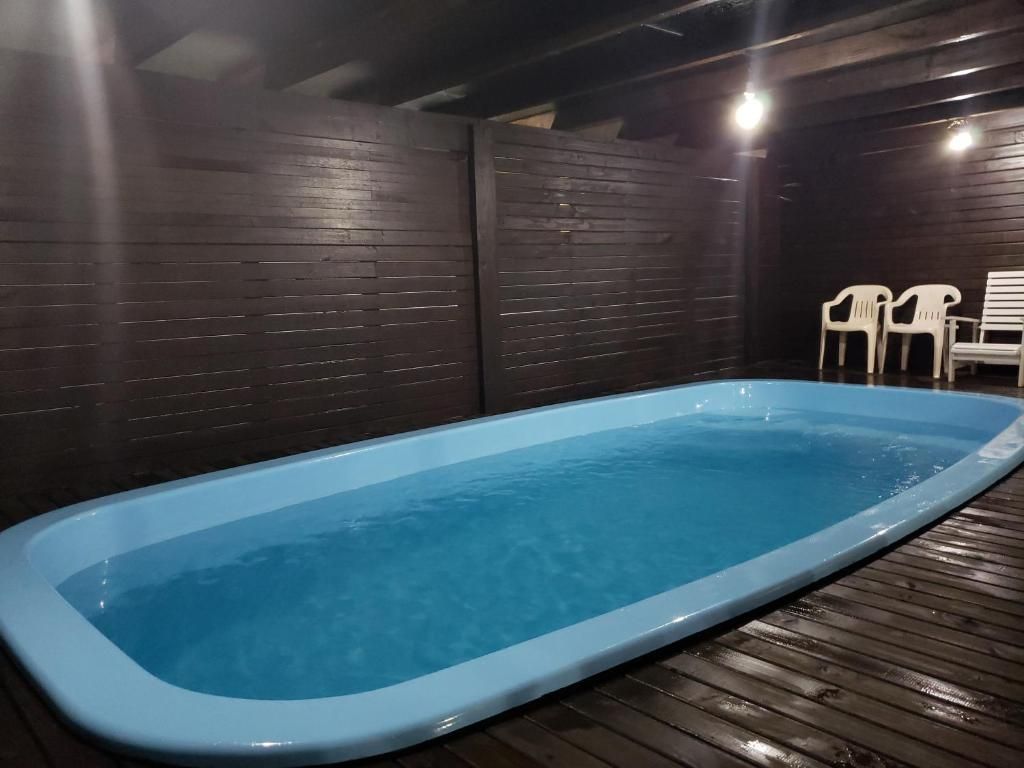 Piscina coberta com água azul, ao lado de um deck de madeira escura e cadeiras brancas no canto