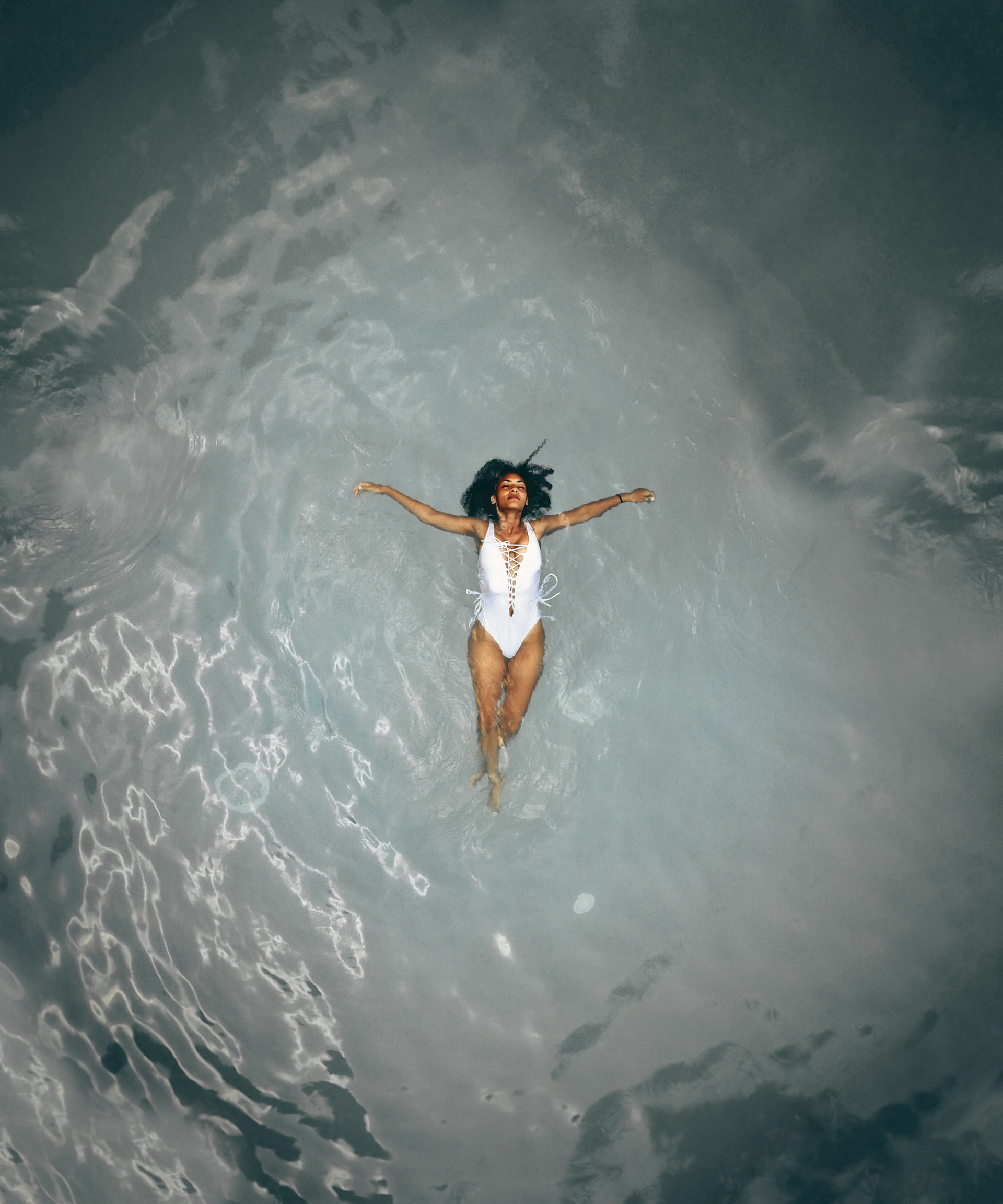 Person floating on back in swirling gray water with arms outstretched, wearing a white swimsuit