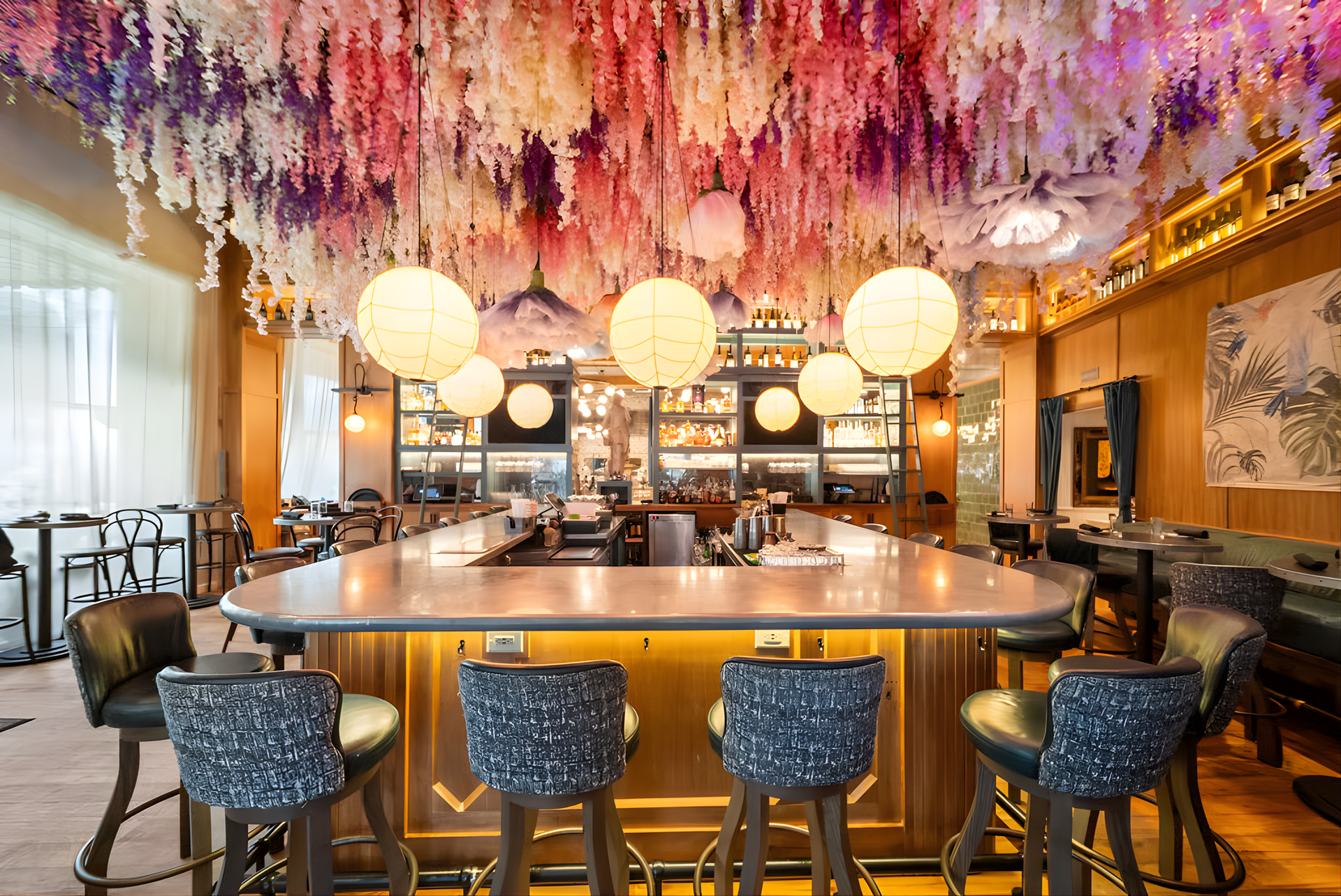 Stylish bar with a curved counter, blue stools, warm lighting, and a ceiling covered in hanging flowers