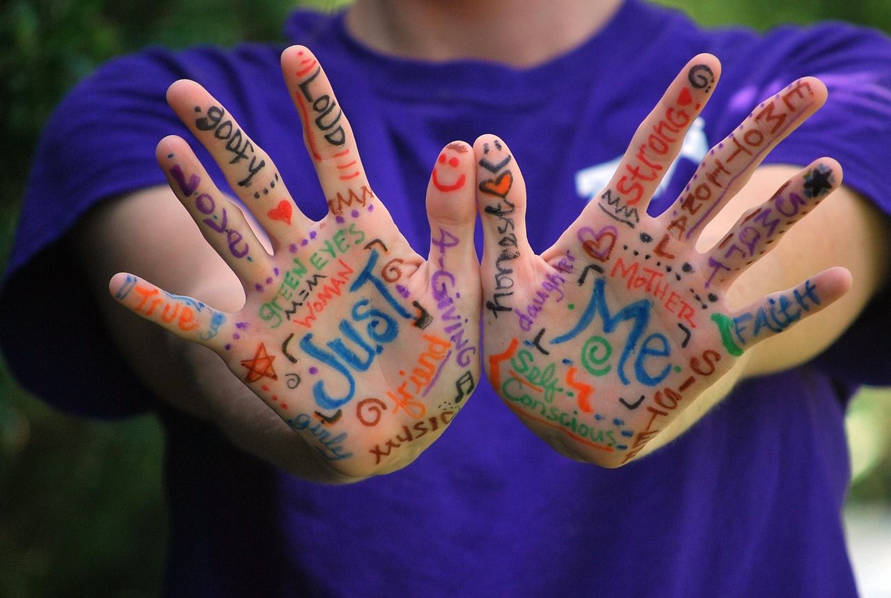 Person holding out hands, each palm written on with colorful words like "Just Me," "Strong," and "Sorry."