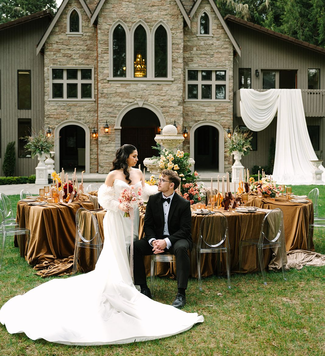 Bride in a white gown holding flowers, standing near a building with a large window and potted plants.