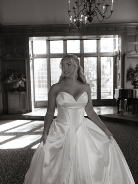 Woman in a strapless gown in a ballroom with large windows and a chandelier.