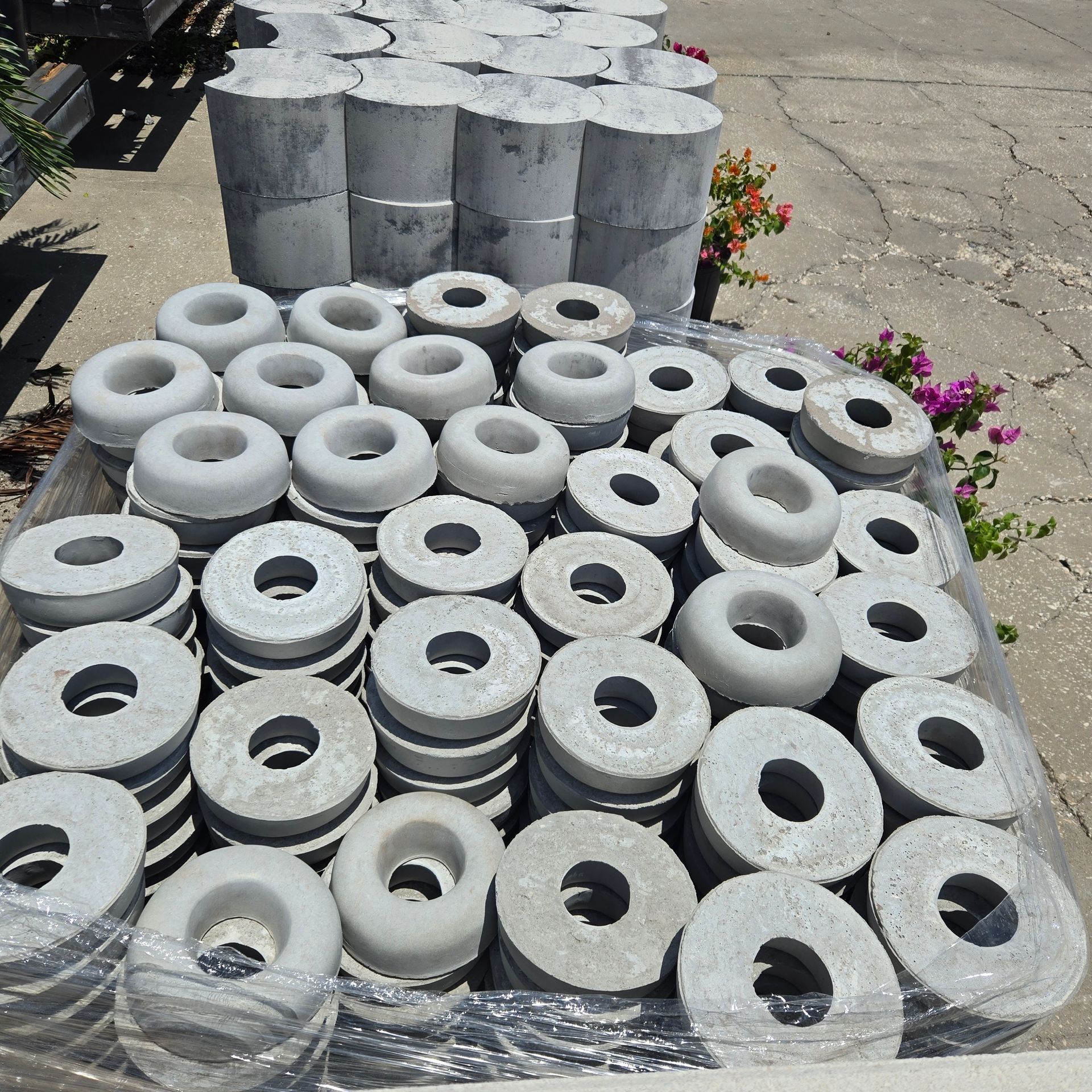 Stacked gray concrete rings on pallets beside a sidewalk, with small flowers in the background.