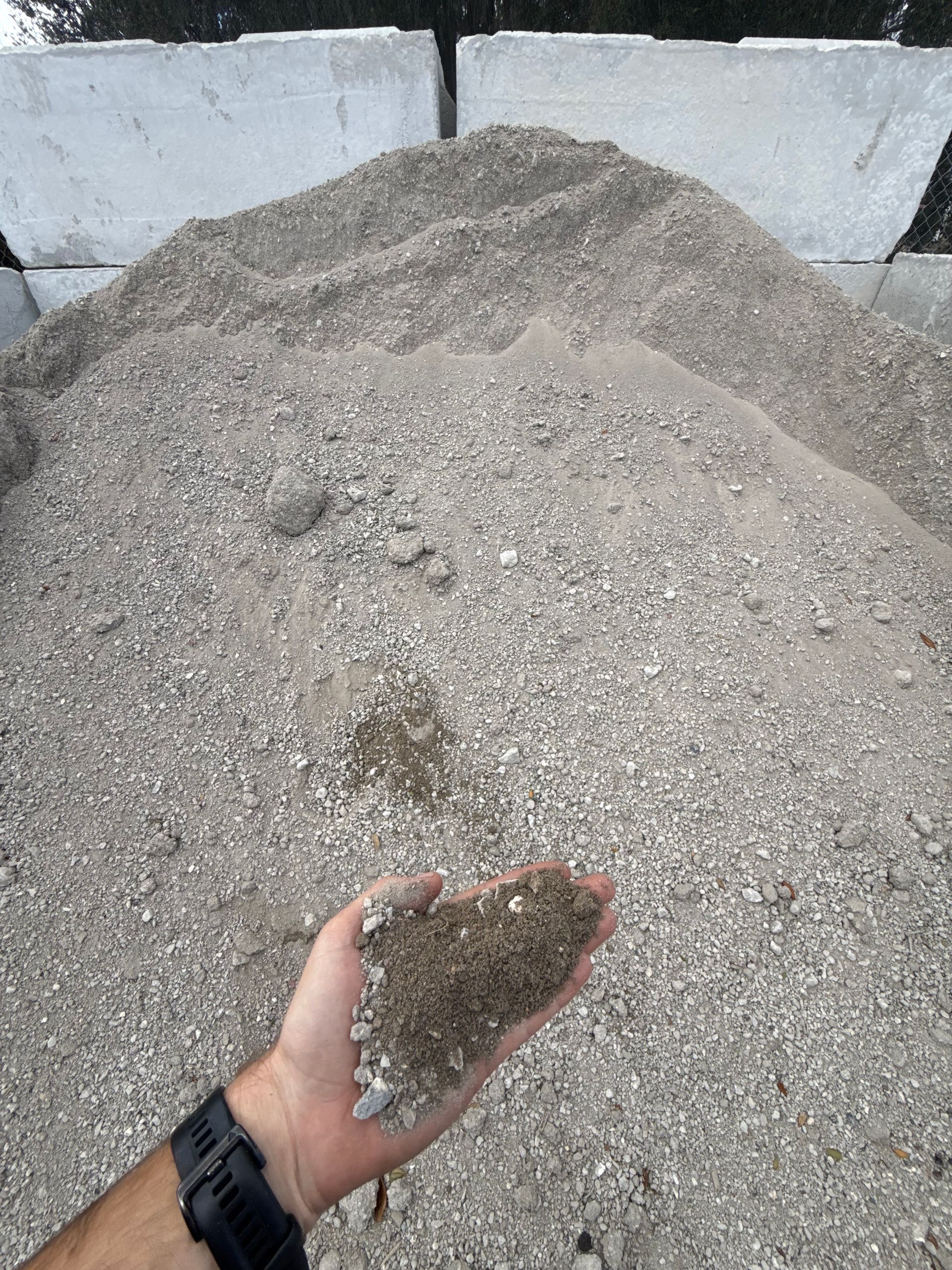Handful of gravel held in front of a large pile of gravel; outdoors.