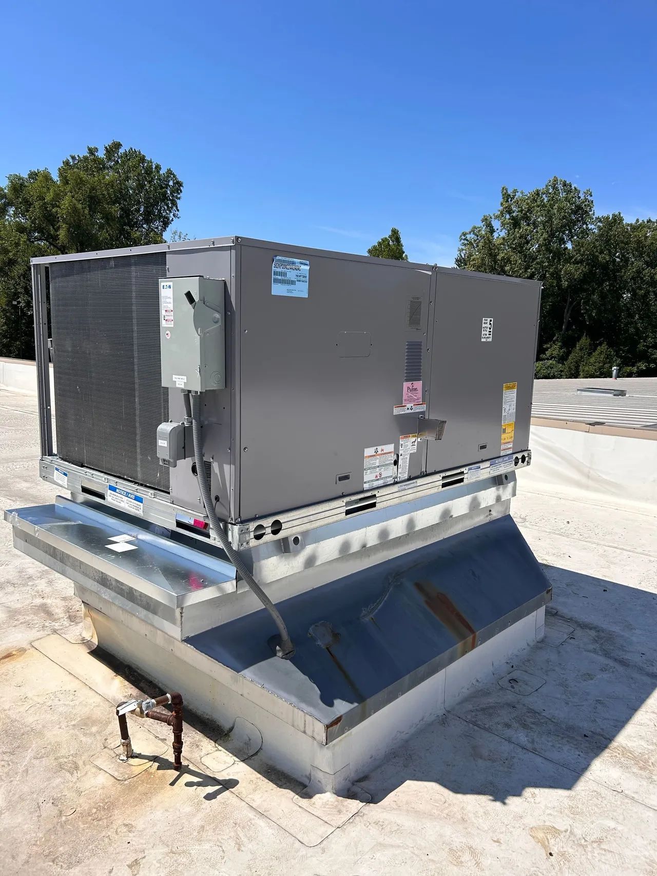 Rooftop HVAC unit on a building, gray metal with a cooling coil, mounted on a raised metal platform, blue sky background.