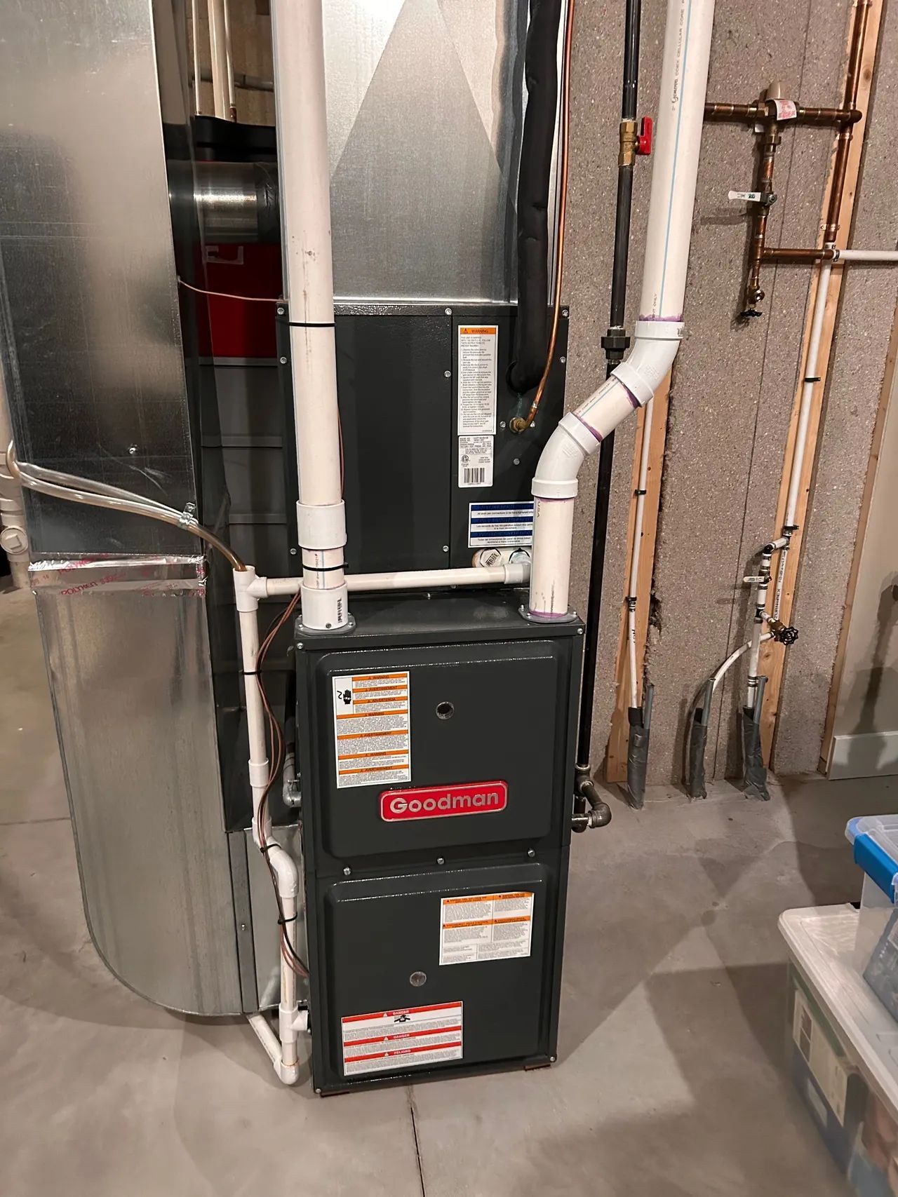Goodman furnace against a wall, with ductwork and PVC pipes, in a basement setting.