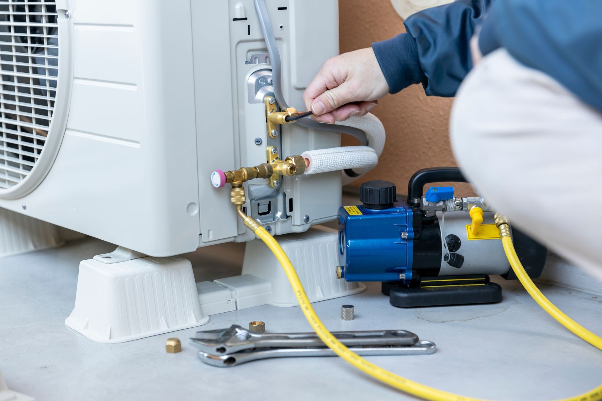 Person servicing an air conditioner outside, using tools and connecting hoses.