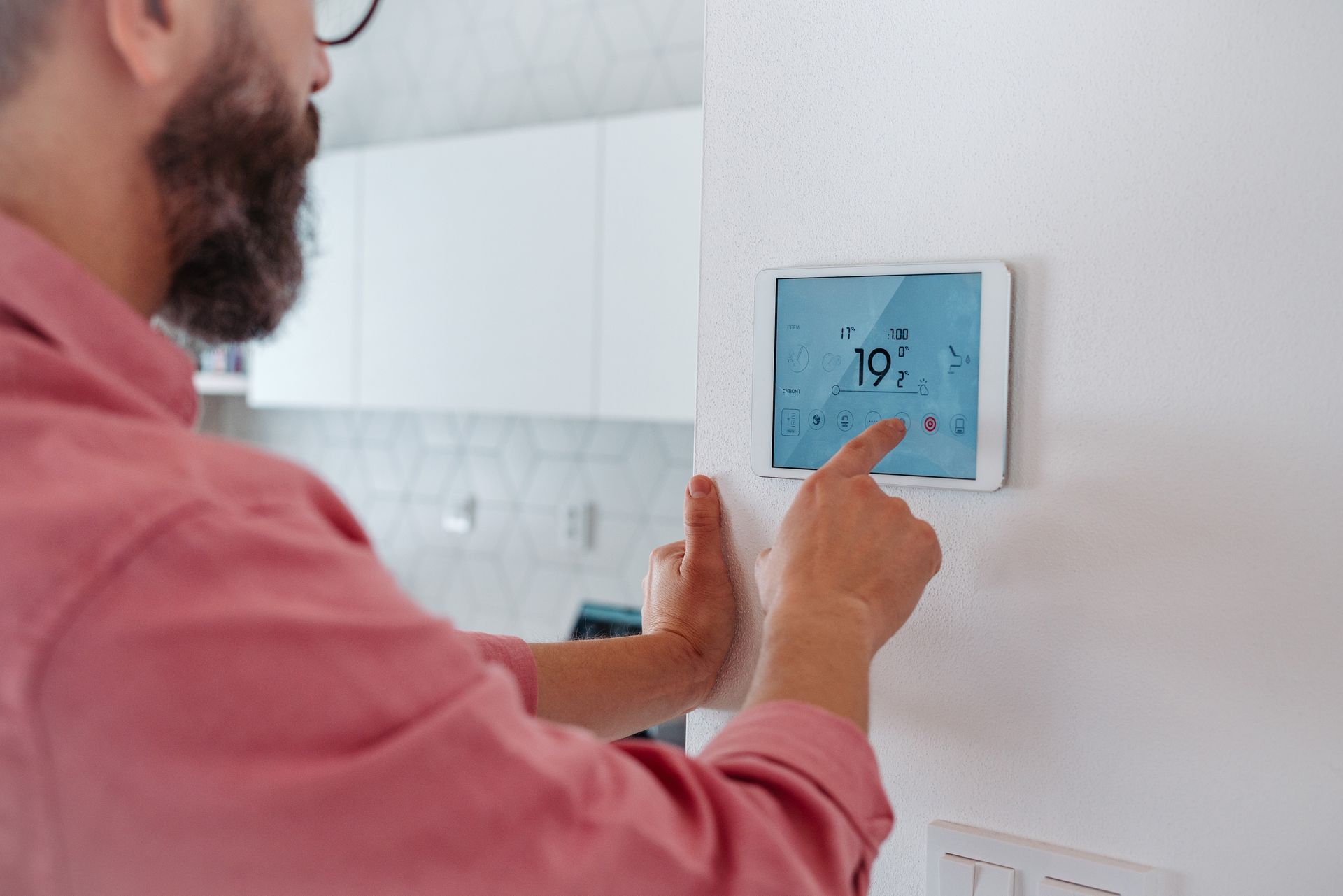 Man in pink shirt adjusts thermostat on white wall in a kitchen.