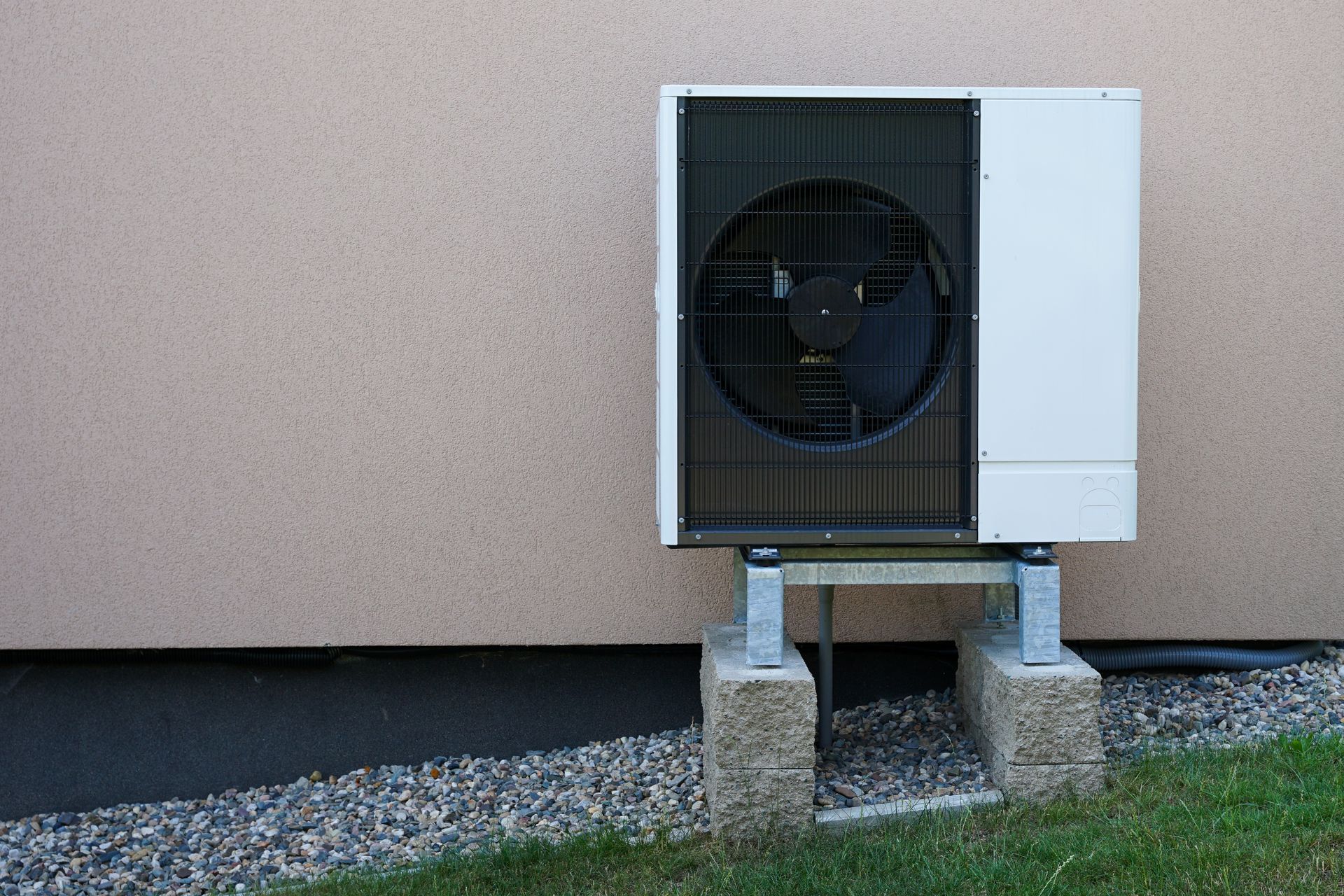 White heat pump unit on a concrete block, mounted outside a building with a tan wall and green grass.